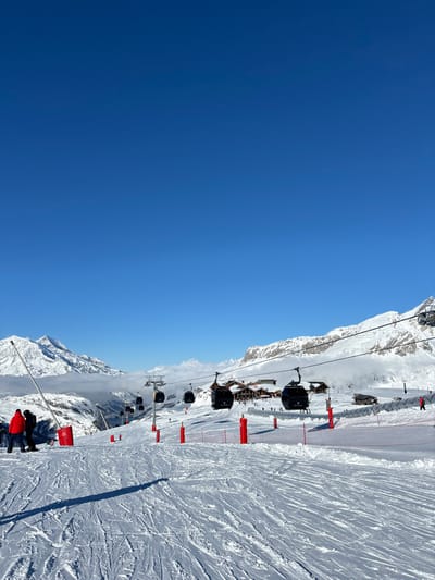 The image depicts a breathtaking, sunny day at a high-altitude ski resort, identified as Val-d'Isère, France. A vast, clear blue sky dominates the left side of the frame, showing a gentle gradient from darker blue at the top to a slightly lighter hue towards the horizon.
On the right, majestic, snow-covered mountains rise dramatically, featuring jagged peaks and rugged terrain. Some clouds are visible clinging to the lower sections of the mountains in the distance. A wide, well-groomed ski slope descends along the right side, crisscrossed with numerous ski tracks from recent activity.
Several black gondola cabins are seen in a line, ascending the mountain via a ski lift system, supported by metal towers. Bright red safety poles or barriers line the edge of the ski slope, providing visual guidance and protection. In the bottom right corner, a few individuals are present on the snow, including one person distinctly clad in a red jacket, indicating skiers or snowboarders enjoying the day.
The bright sunlight suggests it is mid-morning or early afternoon, casting clear shadows on the pristine snow. The scene conveys a sense of exhilarating winter sports and the stunning beauty of a clear day in the French Alps. There is no visible text in the image.