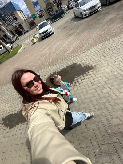 A photograph captures a moment on a sidewalk, likely in Proletarskii, Russia, indicated by a sign in Cyrillic partially visible on a building in the background.
The primary subject is a woman taking a selfie, her face close to the camera, wearing sunglasses and a light-colored jacket. Her long, reddish-brown hair is visible. Behind her, a child lies on the sidewalk, also facing upwards, with what appears to be a scarf or wrap around their head. The sidewalk is paved with gray rectangular bricks, and a dark, irregular stain is visible on the bricks nearby.
In the background, there are several parked cars, including white and dark-colored vehicles. Buildings are visible on the opposite side of the street, one with a sign in Cyrillic. A small patch of green suggests a strip of vegetation alongside the road. The photo is taken at an angle, with the top of the buildings tilted to the left.