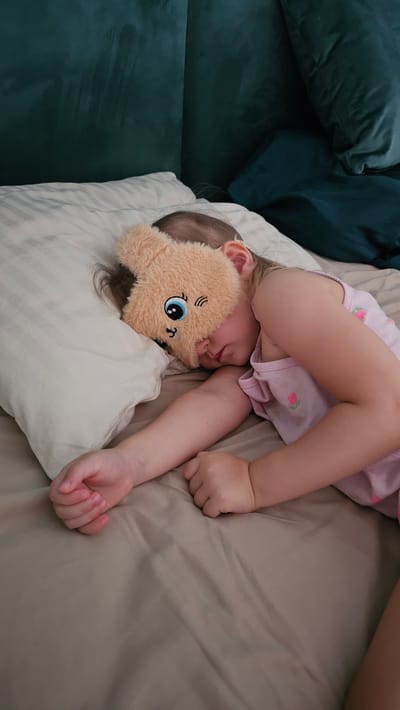 A young child, appearing to be a toddler, is sleeping soundly on a bed. The child is lying on their side, with their head resting on a white pillow. A beige, plush toy shaped like a bear's head is placed over the child's eyes, obscuring their face. The child is wearing a light pink t-shirt adorned with small floral patterns. One arm is bent and resting near their head, while the other arm is extended along their body. The bedding is a neutral, light brown color, and a darker teal couch or large pillows are visible on the left side of the frame, suggesting the child might be sleeping on a couch or in a bed adjacent to one. The lighting suggests it could be daytime, possibly in the afternoon, as there are no strong shadows indicating nighttime. The overall scene conveys a sense of peace and rest. No visible text is present in the image.