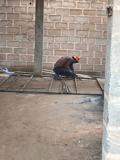 In Okinano, Nigeria, a shirtless individual wearing an orange hard hat and dark pants is crouched on the ground, welding metal rods. The scene is indoors or under a partial structure, with a brick wall behind the person and a concrete floor. A metal frame, likely a gate or fence, is being constructed on the ground, with various metal pieces and welding equipment present. To the right, a large concrete pillar is visible. The environment appears to be a construction site, characterized by unfinished building materials and the presence of manual labor.