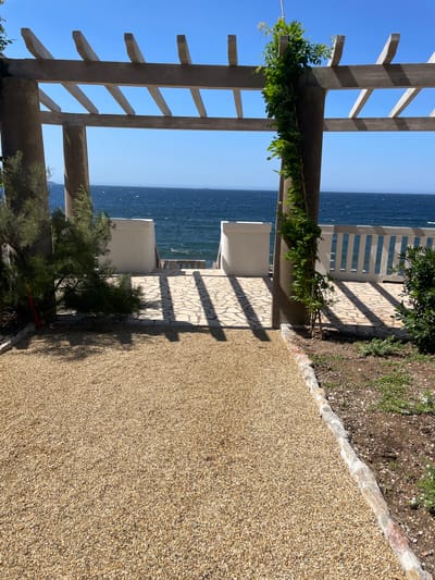 This is a photograph taken outdoors in Carqueiranne, France. The image shows a view through a pergola structure with concrete pillars and beams. The pergola frames a clear blue sky and a body of water, likely the sea, which appears choppy. In the foreground, a path covered in small, light-colored gravel leads towards the water. To the sides of the path are some low-lying green shrubs and a larger, sparse bush on the left. The paved area beyond the gravel path is made of irregular stone slabs, casting distinct shadows from the pergola. There are white, low walls on either side of a set of steps leading down to a white railing overlooking the water. A vine with green leaves is growing up one of the concrete pillars on the right. The overall scene depicts a sunny day with a serene, coastal atmosphere.