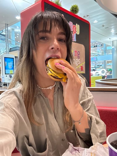 A woman with fair skin, dark hair, and a nose piercing is eating a burger in a McDonald's restaurant. She is wearing a light olive green shirt and a pearl necklace. She also has tattoos on her left forearm and is wearing a silver bracelet. A cup is visible to the upper right of her, and a McDonald's ordering kiosk is visible to the lower left of her. The background shows the restaurant's interior and exterior, consistent with the location of a McDonald's in Spreitenbach, Switzerland.