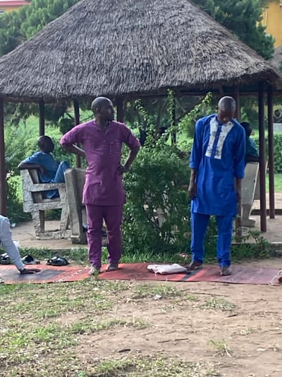 Under a thatched-roof structure, three individuals, presumably men, are outdoors. One is standing erect in a blue ensemble, a white garment rests nearby. Another, clad in purple, leans with a hand on their hip. A third person sits on a rustic, stone-like bench, dressed in blue. The backdrop is a blend of greenery, dirt, and sparse grass.
