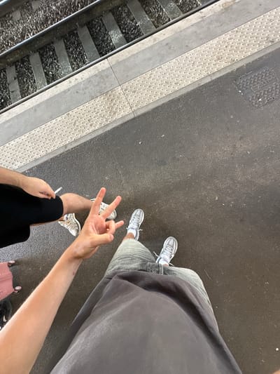 This overhead shot captures two individuals standing on a train platform in Nîmes, France. The scene is set with a train track on the left, a textured yellow safety strip, and a dark asphalt platform on the right. One person is wearing a dark grey t-shirt, light blue jeans, and white sneakers, with their legs and feet visible. They are holding a cigarette in their right hand, and their left hand is extended, displaying a peace sign with their fingers. The other person, partially visible, is wearing a dark grey t-shirt, light grey shorts, and white sneakers. Their legs and feet are also visible. Their right hand is holding a pink suitcase. The lighting suggests it is daytime, and the overall atmosphere appears casual and relaxed. There is no visible text in the image.
