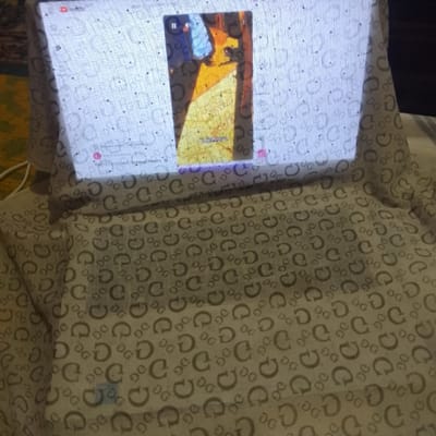 This image shows a close-up view of a digital display, likely a tablet or smartphone, projecting an image onto a white fabric with a repeating pattern of "G" logos. The fabric is draped over an object, creating a makeshift screen. The projected image itself is a still from a video, featuring a brightly lit, abstract scene with yellow and orange tones, and some blue elements. A small, partially visible portion of the screen's interface is also visible, including a YouTube logo in the top left corner and what appears to be a title or text in Cyrillic script. The overall impression is one of creative improvisation, possibly for entertainment or display purposes, set against a backdrop of patterned fabric. There are no people visible in the frame. The lighting suggests it's an indoor setting, potentially during the day or in a well-lit room, as the projection is bright and clear. The location context of Almaluu, Kyrgyzstan, is provided but not directly inferable from the visual content of the image itself.