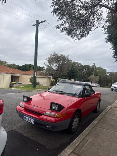 The image shows a red car parked on a street in a residential area. The license plate reads "1BCR-133," which indicates that the vehicle is registered in Western Australia. The car is parked next to a curb with a telephone pole nearby. The sky is overcast, and the trees suggest that the scene is located in a suburb. Based on the license plate, the location is likely within the state of Western Australia, but without additional information or visual cues specific to the area, it is difficult to conclusively pinpoint the scene as Bassendean.