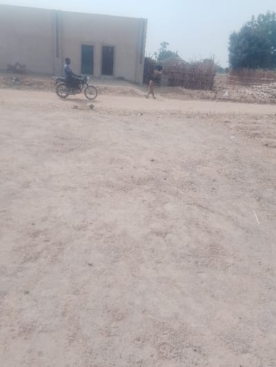 This image depicts an unpaved, dusty environment in Kanwa, Nigeria, under daylight conditions. The foreground consists of light brown, coarse dirt with scattered small stones and sparse vegetation.
In the mid-ground, a dark-colored motorcycle is positioned, facing right. A male individual, wearing a dark shirt and trousers, is seated on the motorcycle. To the right of the motorcycle, a child, dressed in light-colored clothing, is seen walking from left to right.
In the background, on the left, a light-colored, single-story building with two dark, rectangular window or door openings is visible. Further to the right in the background, there is a circular or semi-circular structure constructed from woven natural materials, possibly straw or sticks, surrounded by similar woven fencing. Several trees with green foliage are present in the far background, along with additional areas of dry, sparse vegetation and more unpaved ground. The sky appears light and clear.