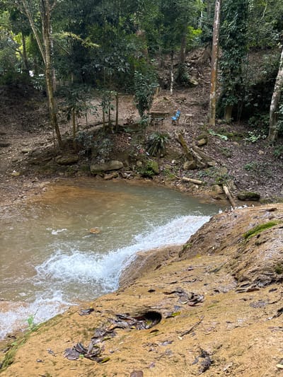 A waterfall cascades into a pool surrounded by lush greenery. The left side features trees and foliage, while the right shows a close-up of a rock face. The water is clear, with visible ripples and movement. Litter is apparent, with a blue plastic bag among other waste items. A picnic table is on the left in the distance.
