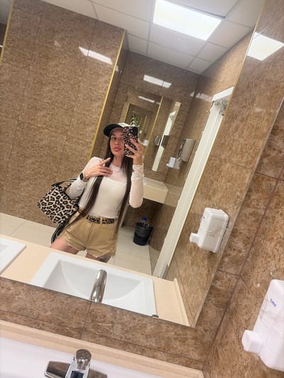 A young woman with long dark hair, wearing a white long-sleeved top, tan shorts, a leopard print belt, and a dark baseball cap, is taking a mirror selfie in a public restroom. She is holding a leopard print phone case and a leopard print purse. The restroom has tan marble tile walls, a white ceiling with fluorescent lighting, and a vanity with two sinks and chrome faucets. There are also soap dispensers and a trash can visible in the reflection. The image appears to be taken in Novosergievka, Russia.