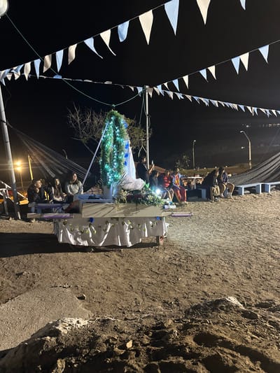 People gather along a street lit by electric lights at night. Pennant banners hang overhead. A structure adorned with lights and flowers sits atop a table draped in white fabric. People are either seated on a platform or standing near the display. The scene takes place next to a rough dirt surface, possibly a sidewalk or roadside.