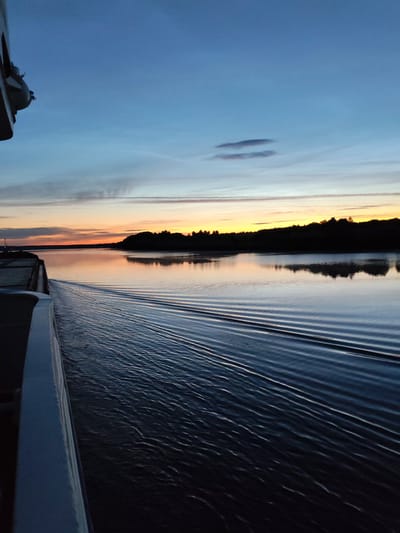 The image captures a serene water scene at dusk, likely in the vicinity of Yakunina Gora, Russia. The sky displays a gradient of colors, transitioning from a deep blue at the top to hues of orange and yellow near the horizon, indicative of a sunset or sunrise. Wispy clouds are scattered across the sky. A dark, silhouetted treeline forms the distant shore. The foreground is dominated by calm water, reflecting the sky's colors, with distinct V-shaped wake patterns emanating from the viewer's perspective, suggesting movement through the water, possibly from a boat. The left side of the image shows the edge of a white structure, possibly a railing or deck of a vessel, extending into the frame. The overall atmosphere is tranquil and evocative of the early morning or late evening hours. There are no people or animals visible in the frame.