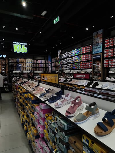 The image depicts the interior of a Skechers store, specifically the "Kids" section, indicated by a large neon sign. The store is well-lit with overhead lighting and displays rows of shoe boxes stacked high on shelves. In the foreground, a variety of Skechers sandals for women are displayed on a white shelf. These include white and pink platform sandals, pink and white heeled sandals with ankle straps, olive green espadrille-style sandals, and dark blue criss-cross strap sandals. Below these displayed shoes, stacks of shoe boxes are visible, many with the "Slip-ins" branding. The store appears to be in a commercial area, with signage for "DONNA" (women) visible on a shelf in the middle ground. The overall impression is of a retail environment designed for shoe sales, with an emphasis on organization and product display. The location is identified as Settimo Torinese, Italy.
