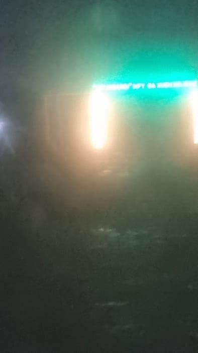 The image depicts a low-light, hazy scene captured at night in Kogon City, Uzbekistan. A prominent, horizontally oriented illuminated sign, emitting a bright green light, dominates the upper-middle portion of the frame. Indistinct white text is visible on this green sign, including characters that appear to spell "MFT OA KOGON." Below and adjacent to this sign, two intense, vertically elongated light sources, characterized by a yellow-orange hue, are present, likely originating from streetlights or building fixtures. A fainter, bluish-white light source is visible on the far left. The environment below the light sources is dark and indistinct, appearing to be a ground surface, possibly wet, which reflects diffuse light. No individuals, vehicles, or specific actions are discernible within the frame.