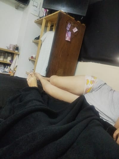 The image depicts an indoor scene, likely a bedroom or living space, with a person relaxing on a bed, largely covered by a black blanket. Only the person's bare feet, legs, and a portion of their torso in a grey t-shirt are visible. They are wearing white underwear adorned with a cartoonish pattern of yellow and brown objects.

The room features white walls and several wooden storage units. To the left, a multi-tiered wooden shelf unit holds various items, including glass jars or containers, a pink appliance (possibly a blender or mixer), and other household goods. A package marked "MAR" is visible on one of its lower shelves. Above this unit, a white router with two antennas is mounted on the wall, clearly displaying the text "Cantv" and partially "Tecnologia", indicating its origin as a Venezuelan telecommunications product.

On the right side of the frame, a taller wooden cabinet or shelving unit has a flat-screen television placed on top. A white folded item, possibly clothing or a towel, hangs from one of its shelves. Attached to the side of this unit is a wire heart-shaped frame, from which several small photographs or cards are clipped. Another flat-screen TV is mounted high on the wall in the upper left corner of the room.

The overall lighting is dim, suggesting the time of day is likely evening or night, or the room is intentionally darkened. The scene portrays a casual, domestic environment, characteristic of a home in Catia La Mar, Venezuela.
