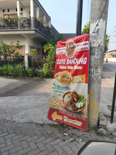The image shows a red banner for "Soto Bandung" placed on the sidewalk in front of a building and a utility pole. The banner advertises the restaurant "KEDAI SI NENG" which is open from 6 AM to 12 AM. It also features images of two dishes, Soto Bandung and Nasi Daun Teruk Ayam Gepuk, with prices 10K and 15K respectively. The banner also indicates that coffee and iced tea are available 10 meters away. The utility pole has several stickers on it, some of which are faded and illegible. The background shows a two-story building with balconies and plants, and a street with a person on a motorcycle in the distance. The ground is paved with concrete and cobblestones. The scene is set in the city of Barengkrajan, Indonesia.