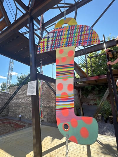 An outdoor, vertical, colorful, stylized art installation, resembling a mushroom or a sock, hangs from a metal structure under a bright blue sky with scattered clouds. The installation features a patterned top with plaid-like designs in various bright colors, a long stem with bands of multi-colored stripes and large orange polka dots, and a bulbous, green base adorned with orange polka dots. A circular cutout is visible on the lower part of the base. The art piece is suspended by metal chains and hooks. In the background, a brick wall is visible, along with a wooden deck, an ornate rug on the ground, and some greenery. To the right, a person's hands are seen reaching towards the installation, holding what appears to be a phone or camera. To the left, a white sign with text is attached to a black metal support post. The overall scene suggests an art exhibition or public display in an urban outdoor setting. This artwork is located at the Yelets Museum of Local Lore in the city of Yelets, Russia.