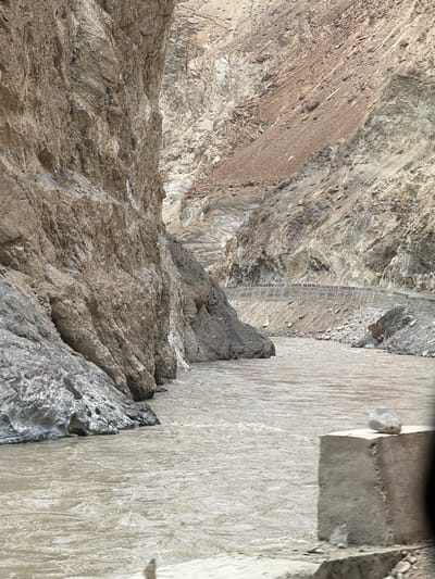 A wide, muddy river flows through a narrow canyon. Steep, rocky cliffs rise on either side of the river, with a road visible carved into the side of the right cliff. The terrain is arid and mountainous, with sparse vegetation. The water appears to be fast-moving. The scene suggests a remote, natural landscape. This location is in Stongde, India.