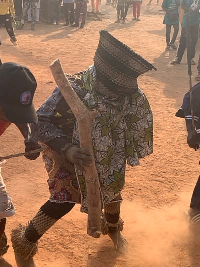 A person adorned in a traditional, patterned outfit and a distinctive woven headdress is captured holding a thick branch. This individual appears to be part of a cultural event or performance, as suggested by the presence of a crowd in the background. The ground is dusty, and the atmosphere is filled with a sense of activity and movement. Additional people are visible around the main subject, and a person wearing a baseball cap is partially visible in the lower portion of the frame.