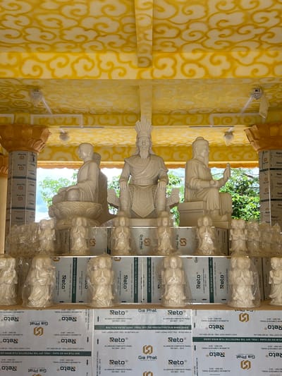 The image depicts an open-air shrine or temple structure located in Đăk Mar, Vietnam. The ceiling is painted yellow with a repeating white cloud motif and features several white ceiling fans. Ornate, gold-colored pillars with lotus-like capitals, partially covered by protective film, frame the scene on the left and right.
Three large, light-colored carved stone statues are positioned centrally. On the left, a seated figure with a bald head and monastic robes meditates on a lotus pedestal. In the center, a prominent, bearded figure with an ornate headdress and ceremonial attire is seated on a throne. To the right, another bearded, robed figure with a hat sits with one hand raised.
Below these main statues, multiple rows of smaller, light-colored carved figures, many of which are individually wrapped in clear plastic, are displayed. These figures typically show individuals with hands clasped. They are arranged on platforms constructed from stacked rectangular panels. These panels are silver-colored aluminum composite panels, branded "Gia Phat" and "Reto," with text in both Vietnamese and English indicating "ALUMINIUM COMPOSITE PANEL" and instructions regarding protective film removal. Green foliage and trees are visible in the background, suggesting an outdoor or semi-outdoor setting.