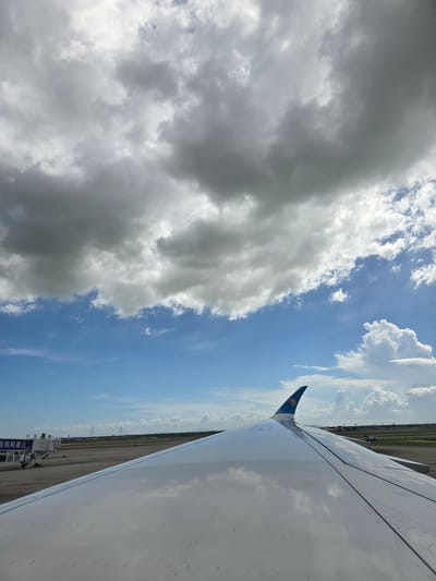 The image shows a view from an airplane window, looking towards the wing and the sky. The wing is white and reflects the clouds above. The sky is blue with fluffy white clouds, and some darker storm clouds. In the distance, an airport tarmac is visible with a jet bridge and some airport buildings. The location is Bao'an District, China, which is home to Shenzhen Bao'an International Airport. The scene appears to be at an airport, possibly before takeoff or after landing.