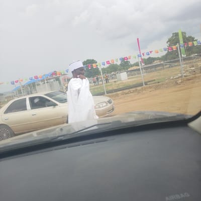 In the city of Wuro Ngangandi, Nigeria, a man dressed in traditional white attire and a white headwrap stands beside a beige sedan. The man is positioned to the right of the vehicle, facing left, with his left hand touching his chin. The sedan, appearing to be a Toyota Camry, is parked on a dirt road. In the background, a chain-link fence encloses a construction site with various structures, including what appear to be covered shelters. Several colorful triangular flags on a string are strung across the top of the fence, and a pink flag is visible to the left. Several individuals, some wearing orange vests, are visible working within the fenced area. The sky is overcast, suggesting a cloudy day. The foreground shows a portion of a car's dashboard and windshield, indicating the photo was taken from inside a vehicle.
