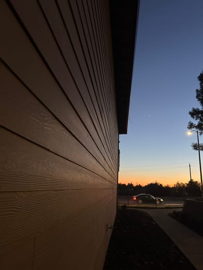 An exterior shot captures a building's wooden siding illuminated by the warm glow of a setting sun. The siding is a warm, light brown color with visible wood grain patterns. The building's roofline, a dark, contrasting color, runs along the top edge of the frame. The sky transitions from a deep blue at the top to a vibrant orange and yellow near the horizon, indicating dusk.

In the distance, a car is visible, its headlights on, traveling along a road. The road is bordered by a fence. A streetlight is on, casting a yellow light. A single bright point of light, possibly a planet or star, is visible in the clear sky. The scene appears to be in a suburban or urban environment, given the presence of the road, car, and streetlight. The overall atmosphere is serene, with the natural beauty of the sunset contrasting with man-made elements.