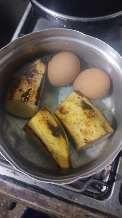 The image shows a silver pot sitting on a stovetop. Inside the pot are two brown eggs and four pieces of peeled plantain submerged in water. The stove appears old and slightly dirty. The location of Putucual, Venezuela is not evident in the image.