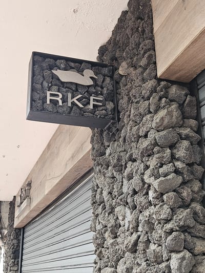 A low-angle, outdoor shot depicts architectural details of a building, potentially in the city of Osorno, Chile. The focus is on the exterior wall and ceiling intersection. The wall is painted a light off-white, while the ceiling features exposed wooden beams and corrugated metal. A decorative stone facade is seen along the edge of the ceiling. A square sign, filled with similar rocks, hangs from the ceiling. The sign contains a metal silhouette of a bird-like figure, the letters "R," "K," and "F."