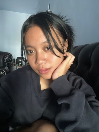 A young female with dark hair is captured in a close-up, selfie-style portrait. She is leaning her right cheek on her right hand, which is adorned with a simple ring on her ring finger. Her hair is somewhat disheveled, with strands falling across her face and forehead. She is wearing a dark, crew-neck sweatshirt. The background features a dark, upholstered couch on the right and a light blue wall on the left, with some shelves and objects visible on the wall. The scene appears to be indoors, possibly a living room. The lighting is dim, suggesting it might be evening or a low-light indoor environment. The woman's expression is neutral to slightly pensive. The image quality is moderate, with some visible texture and grain.
