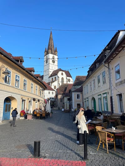 The image shows a street view in Sibiu, Romania, dominated by the Council Tower. The street is paved with gray cobblestones and has buildings on both sides, some with outdoor cafe seating. A woman in a light-colored jacket is leaning against a black post. Other people are present in the cafes and walking in the street. The sky is clear and blue. Powerlines stretch across the sky.