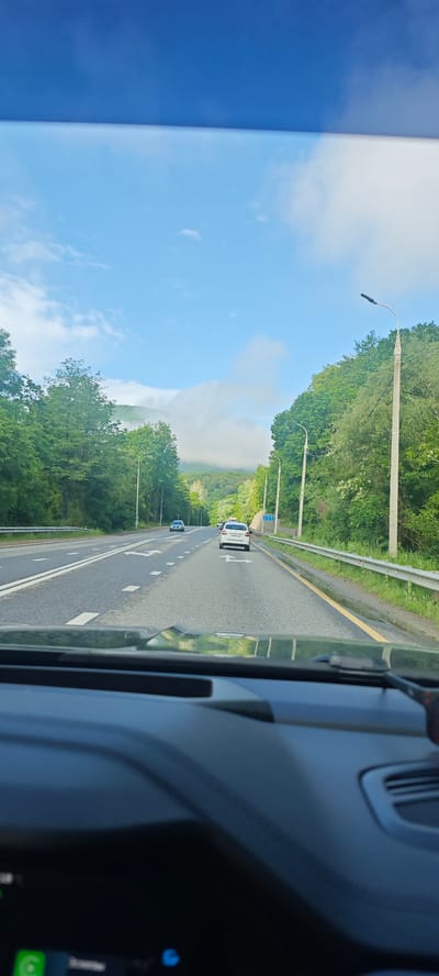 The image depicts a view from inside a car driving on a multi-lane road. Two cars are visible ahead, one white and the other black. Road markings indicate lane divisions and directional arrows. Trees line both sides of the road, and in the distance, mountains are partially obscured by clouds. The sky is blue with scattered clouds. Streetlights are present along the roadside. The lower portion of the image shows the car's dashboard and windshield. The location is Moldavanovka, Russia.