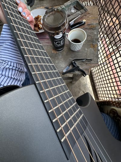 The image depicts a close-up view of a black guitar with a fretted neck, positioned diagonally across the frame. A person wearing a blue and white striped shirt is holding the guitar.

In the upper left corner, a weathered wooden surface holds several items, including a white ceramic cup with coffee residue, a black can of Landessa Espresso Ice Coffee, an open glass jar containing a dark liquid, a small plate with food, a black lighter, and a black capo. A metal mesh structure is visible in the upper portion of the image, providing a backdrop to the scene.

The setting suggests an outdoor location, possibly a patio or garden area. The image was most likely taken somewhere other than Agarak, Armenia, since Landessa Espresso Ice Coffee is not widely available in Armenia.