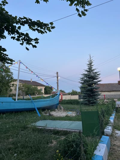 The image depicts an outdoor scene in the city of Slanchevo, Bulgaria, during twilight. In the foreground, a vibrant blue, wooden boat is prominently displayed on land, with an oar resting inside. The boat is adorned with a string of colorful triangular flags that stretch across the frame. Behind the boat, a tall, mature evergreen tree stands to the right, and various plants and flowers are visible. A green metal gate is positioned to the right of the tree, with a decorative border of white and blue concrete blocks in the lower right corner. Several residential buildings with tiled roofs are visible in the background, along with utility poles and power lines. The sky is a soft gradient of blue and pink, indicating the transition from day to night. No people are present in the scene.