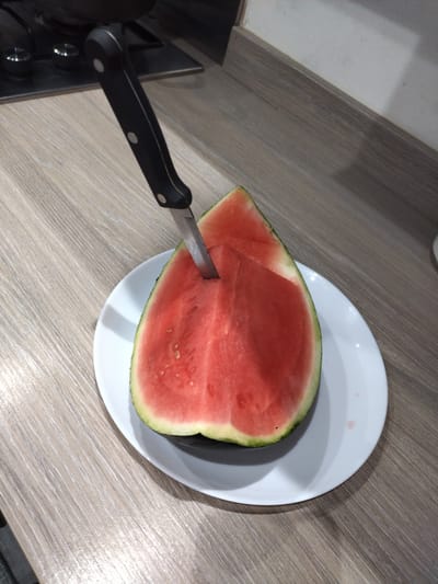 A slice of watermelon is presented on a white plate, with a black-handled knife inserted into it. The watermelon is a vibrant red, with visible seeds and a green rind. The plate rests on a light-colored wooden countertop. In the background, a stovetop is partially visible, indicating a kitchen environment. The lighting suggests an indoor setting, possibly during the day or with artificial light. The image does not provide specific clues to determine its exact location in Darlington, United Kingdom.