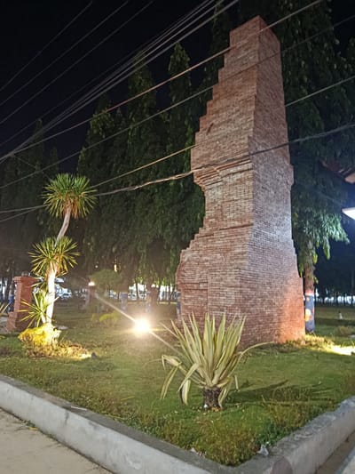 This media file shows an outdoor scene at night, likely in Besuki, Indonesia. The main subject is a tall, brick structure with a stepped, almost fragmented design, resembling a ruin or an incomplete monument. The brickwork is detailed, showing individual bricks and mortar lines. In front of the structure, there is lush green grass and several types of plants, including a spiky, light-green succulent and a palm-like tree with curved trunk and spiky foliage.

Illuminated by bright spotlights, the grass and plants are lit up, casting shadows and creating a dramatic effect. In the background, numerous tall, dark trees form a dense backdrop against the night sky, which is crisscrossed by a network of overhead wires, suggesting an urban or semi-urban environment. The overall mood is somewhat mysterious and serene, enhanced by the nighttime setting and the stark lighting.

There are no people clearly visible in the foreground or midground. In the far background, very small, indistinct figures can be seen on the grass, possibly people walking or relaxing. The lighting suggests it is a public space, perhaps a park or a plaza. The weather appears to be clear, given the nighttime setting and lack of rain. The brick structure, along with the tropical-looking plants, might indicate a historical or cultural site. No visible text is present in the image.