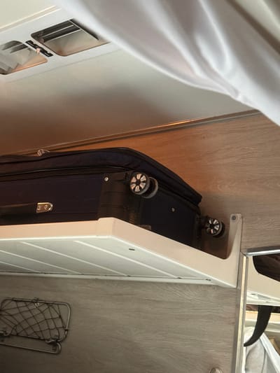 In Gryazi, Russia, an interior scene is depicted focusing on a dark blue, soft-sided wheeled suitcase resting on a white, ribbed overhead shelf. The suitcase, positioned horizontally, displays two of its four wheels, which exhibit a distinctive star-like spoke pattern and visible accumulation of dirt or grime. The white shelf itself is mounted to a light wood-grain patterned wall, secured by a white L-bracket on the right. Below the shelf, a small, black metal mesh storage rack is affixed to the wood-grain wall. Above the suitcase and shelf, a light-colored (likely white) wall or ceiling features two rectangular vents with metallic grilles in the upper left corner. A white, draped fabric, consistent with a curtain, is visible along the right edge and upper right portion of the image. To the far right, a reflective surface, possibly a mirror, partially reveals indistinct dark and light objects. The overall environment suggests a compact travel compartment, such as a train cabin or a recreational vehicle. No individuals are visible.