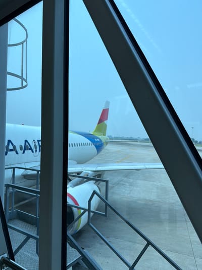 The image depicts an airplane seen through the window of an airport jet bridge. The aircraft is white with blue lettering that appears to read "AAir," and a tail fin with green, blue, red, and white markings. The plane is parked on the tarmac, with the jet bridge extending towards it. A set of stairs with metal railings is visible in the foreground, leading down from the jet bridge. The environment appears to be an airport, with concrete pavement and some distant structures or airport equipment visible. The sky is overcast and pale blue. There are no people visible in the image. The location is identified as Rawa Burung, Indonesia.