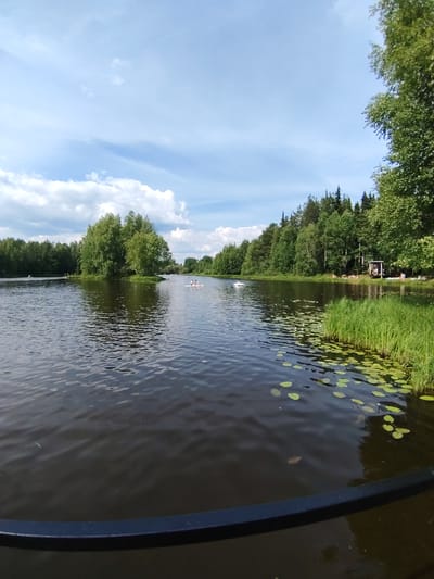 In the city of Vyltydor, Russia, a serene lake scene unfolds under a partially cloudy sky. The foreground features a dark body of water with gentle ripples, reflecting the sky and the surrounding greenery. Lily pads are scattered across the surface of the water, particularly on the right side of the frame.
A small, tree-covered island is visible in the middle distance, adding a natural focal point to the lake. On the water, two small kayaks with people can be seen paddling, suggesting recreational activity. Further in the background, the shoreline is densely lined with various trees, creating a lush, green backdrop.
On the right side of the lake, a wooden structure resembling a small gazebo or covered seating area is present, with a few individuals relaxing nearby. The overall atmosphere is one of peaceful outdoor recreation in a natural setting.