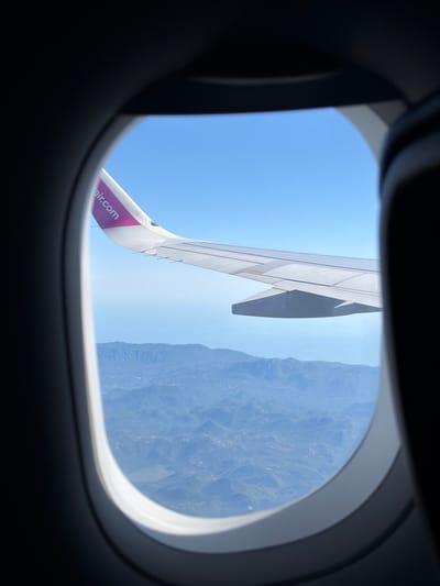 The image shows a view from an airplane window. The window frame is dark, with a bright, oval-shaped opening revealing the sky and landscape below. A portion of an airplane wing, painted white with a magenta logo reading "air.com", extends into the frame from the upper left. The wing is positioned above a vast, mountainous terrain. The mountains appear to be covered in a light green vegetation, with visible valleys and peaks. The sky is clear and a vibrant blue. The scene suggests an aerial view of a rural, possibly coastal, region, consistent with the mountainous landscape of Donja Cijevna, Montenegro.