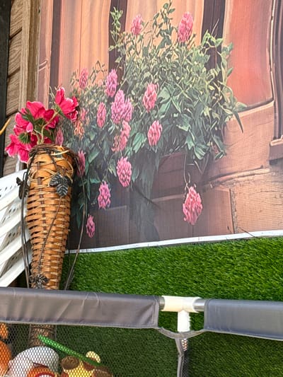 The image shows a close-up of a floral arrangement against a backdrop that appears to be a printed image or canvas depicting a building facade with flowers. The dominant colors are pink, green, brown, and gray.
The floral arrangement consists of pink flowers and green foliage. It is partially situated in front of a woven basket or container.
To the right, a section of what looks like a padded or fabric-covered frame is visible, positioned against an area of artificial green turf.
No people, specific actions, or interactions are evident. The location is not discernible.