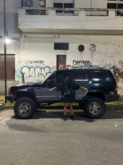 A photograph taken in Florida, Argentina depicts a black SUV, likely a Jeep Cherokee, parked on a street next to a wall heavily covered in graffiti. A person stands next to the SUV, with their arm extended toward the vehicle. The building behind the SUV is multiple stories and shows signs of aging, with exposed pipes and wiring. Street lighting is present, illuminating the scene. The pavement of the road appears uneven. The tires of the SUV are large and appear well-worn. The image overall has a gritty, urban feel.