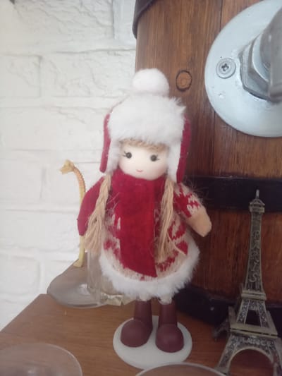 A posed, medium shot shows a doll dressed in winter clothing. The doll has blonde braided hair and is wearing a red and white knitted sweater, a red scarf, and a red hat with a white pom-pom. The doll stands on a white circular base, with maroon boots. To the left of the doll, a yellow dinosaur figurine is partially visible, standing behind a glass object. To the right of the doll, a miniature Eiffel Tower model is placed on the wooden surface. The background consists of a white brick wall and a dark wooden barrel-like object. The scene is indoors, on a wooden shelf.