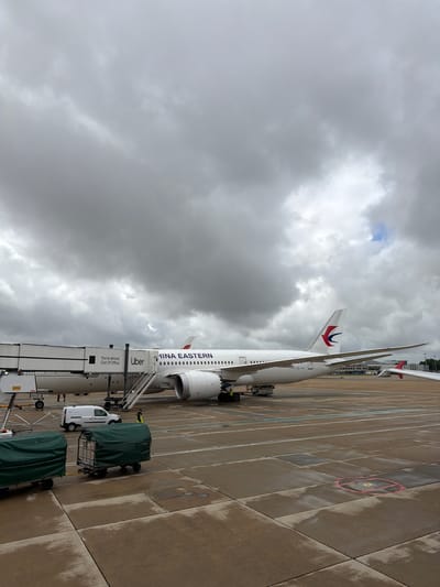 This image captures a China Eastern Airlines Boeing 787 Dreamliner (registration prefix B-) parked at a gate on a wet tarmac under a heavily overcast sky at London Gatwick Airport, near Crawley, United Kingdom. The white aircraft, featuring "CHINA EASTERN" in blue text and the airline's red and blue swallow logo on its tail fin, is connected to a white jet bridge. The jet bridge displays prominent "Uber" branding in black, accompanied by the text "You're almost Out Of Office." A set of mobile stairs extends from the jet bridge to the tarmac. In the foreground, two green tarp-covered baggage carts are parked. A white Renault Kangoo cargo van, a yellow ground power unit, and a DHL-branded piece of ground equipment are visible near the aircraft and jet bridge. At least two airport ground crew personnel, wearing high-visibility vests, are present on the tarmac near the aircraft. The concrete tarmac shows signs of recent rainfall, and the sky is dominated by thick grey clouds, with a small patch of brighter sky visible in the upper right quadrant.