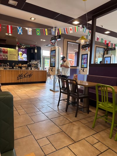The image shows the interior of a McDonald's restaurant in Wolfsburg, Germany. The scene is a typical fast-food establishment with a McCafe counter, digital ordering kiosks, and seating areas. In the foreground, there are two chairs, one dark brown and one bright green, at a wooden table. A purple bench is visible behind the table. The floor is tiled with large, square, beige tiles.
In the mid-ground, a man wearing a white t-shirt and a black baseball cap is standing in front of a digital ordering kiosk, looking at his phone. The McCafe counter is to the left, featuring a menu board with specials and drinks. Behind the counter, coffee machines and other restaurant equipment are visible.
In the background, a string of international flags is hung across the ceiling, suggesting a connection to a sporting event or a celebration of diversity. Digital menu boards are also visible, displaying food and drink options. The overall environment is brightly lit and clean, characteristic of a commercial dining space. The presence of specific McDonald's branding and the German text on the ordering kiosks confirm the location as Wolfsburg, Germany.