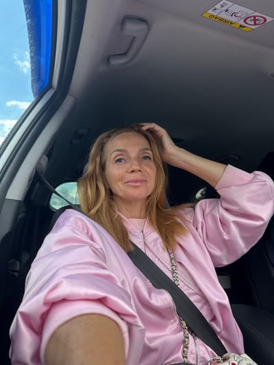 A woman is taking a selfie in a car. She is wearing a pink satin bomber jacket over a pink sweater. Her hair is blonde and falls to her shoulders. She is smiling at the camera. The car's interior is visible, including the airbag warning sticker on the sun visor. The sun visor itself is gray, as are the car's A-pillars. The sky is visible through the car window, and it appears to be a clear day. The time of day is likely daytime. The overall mood of the image is casual and cheerful. There is visible text on the airbag warning sticker which says "AIRBAG".