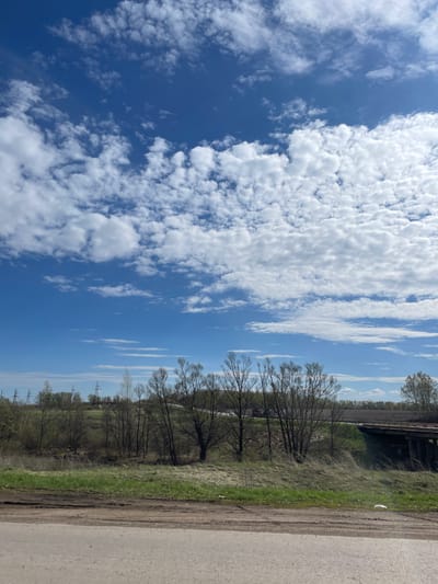 The image shows a landscape in Ulukulevo, Russia. The sky is mostly blue with numerous white cumulus clouds. A road runs along the right side of the image. A green strip of land separates the road from a line of leafless trees. Power lines can be seen in the distance. A bridge is visible in the top right corner.