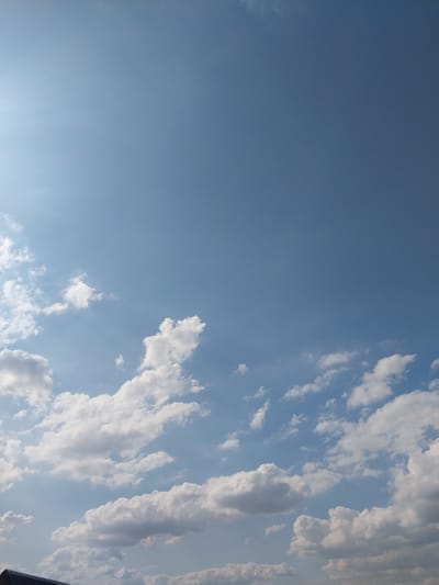 The image shows a clear blue sky with scattered white cumulus clouds. The sun is visible in the upper left corner, casting a slight glare. The bottom of the frame shows a sliver of a dark, sloped structure, possibly a rooftop, suggesting an outdoor scene in Nizhneye Myachkovo, Russia.