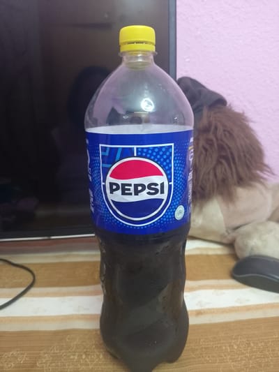 A half-full one-liter bottle of Pepsi, with a yellow cap, stands on a striped surface. The Pepsi logo is clearly visible on the blue label. In the background, a dark surface, possibly a television, is visible, along with a pink wall and a brown stuffed animal with fuzzy fur. To the right of the bottle, a black computer mouse rests on the striped surface. A black cord snakes across the striped surface from the left side of the image. The scene appears to be indoors. There is no information within the image to suggest the location is Urgench, Uzbekistan.