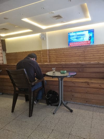 The image shows a man sitting alone at a small table in a cafe in Tunis, Tunisia, at coordinates 36.838, 10.128. He is wearing a dark blue shirt and jeans, and he has his back to the camera. His left hand is resting on the table. A black bag is on the floor next to the table. There is a glass of water on the table. The cafe has wooden paneling on the walls. There is a TV screen showing what appears to be a news broadcast in Arabic. There is a ceiling fan in the corner of the room. The floor is tiled.
