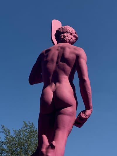 The image shows a pink statue of a muscular, male figure from behind, set against a clear blue sky. The statue has curly hair and is holding something in its clenched fist. Below and to the left of the statue, the tops of green trees are visible. There is no indication that this scene is located in Zhabkino, Russia.